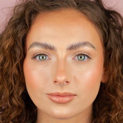 Close-up of a person with curly brown hair, light blue eyes, and a septum piercing looking directly at the camera. With distinct brows and Boldface Lashes - Short and Sweet, they're wearing cruelty-free natural makeup and a soft pink lip color.