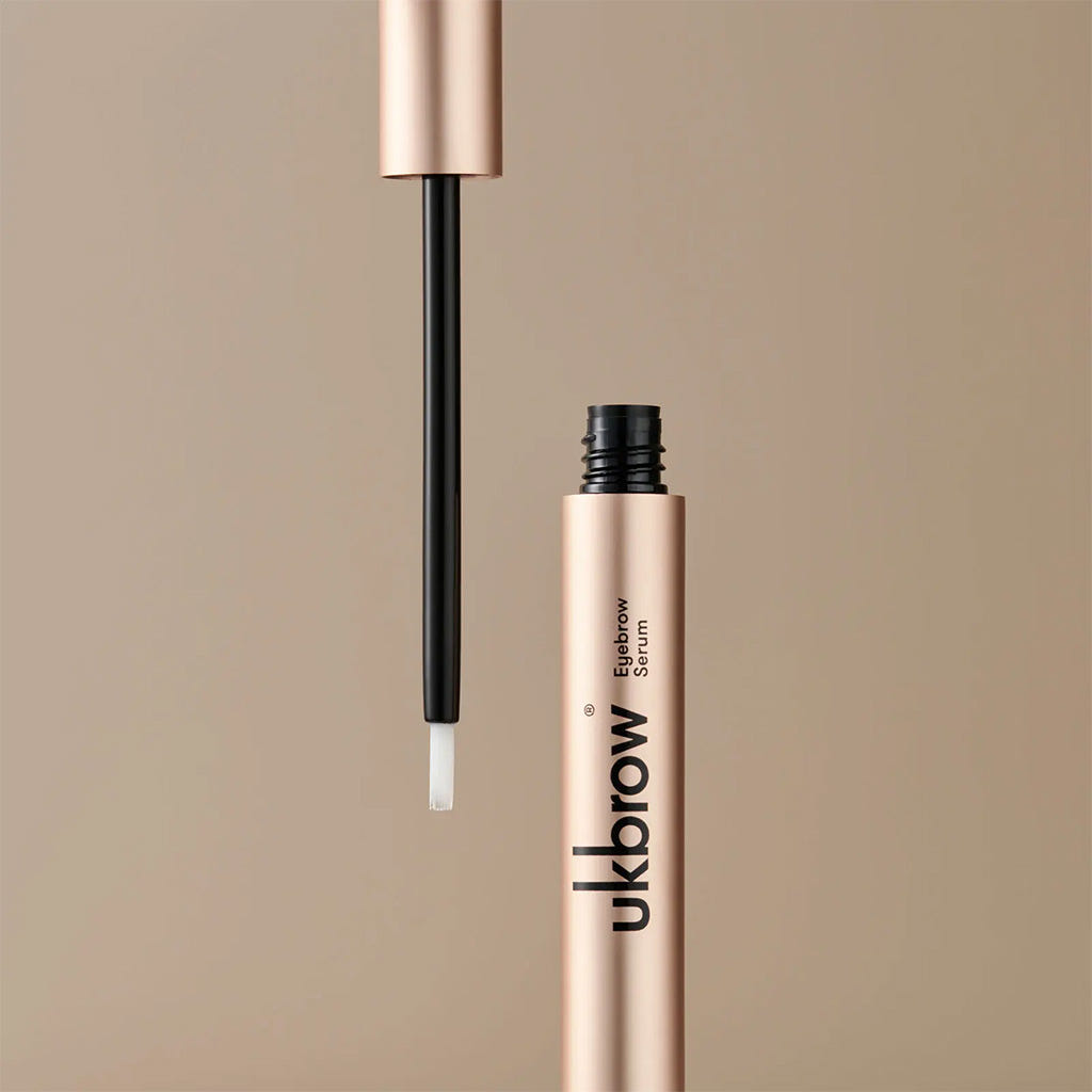A gold tube of UKLASH's UKBROW Eyebrow Serum (3ml), crafted as a powerful enhancer for brow hair growth, sits gracefully with its applicator wand unscrewed and ready for application. The background features a smooth, light beige hue.