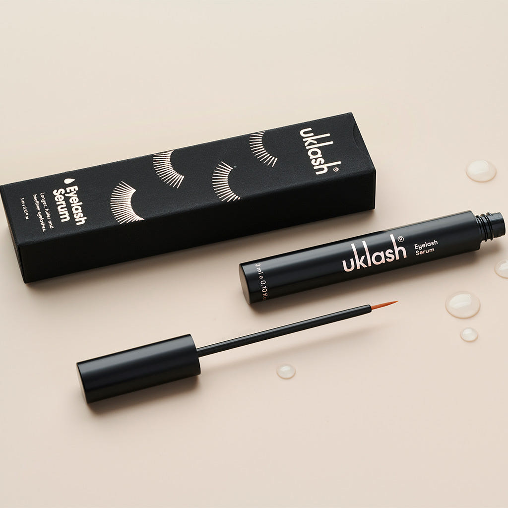 A 3ml black tube of UKLASH Eyelash Serum, renowned for enhancing eyelash length and its cruelty-free formulation, is placed beside its corresponding black box decorated with white eyelash designs. The neutral beige background displays a few dispersed droplets.
