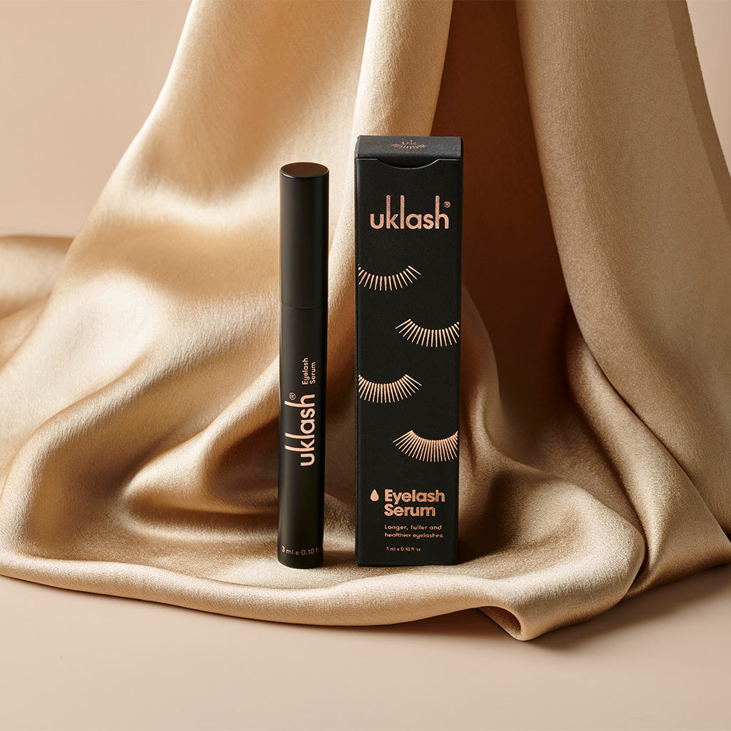 The UKLASH Eyelash Serum (3ml) by UKLASH, known for being cruelty-free and promoting longer lashes, is beautifully displayed alongside its chic black and gold packaging. The box, adorned with an illustration of lush eyelashes, rests elegantly on a backdrop of sumptuous, flowing gold fabric.