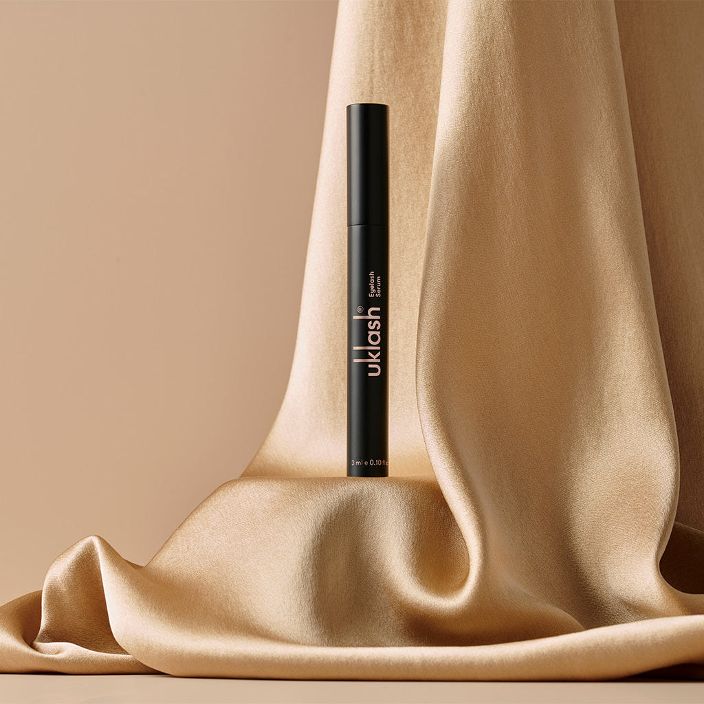 A black tube of the 3ml UKLASH Eyelash Serum stands upright against beige, flowing fabric. The smooth, silky texture creates a luxurious backdrop for this cruelty-free product from UKLASH, promising longer eyelashes with its sleek design.