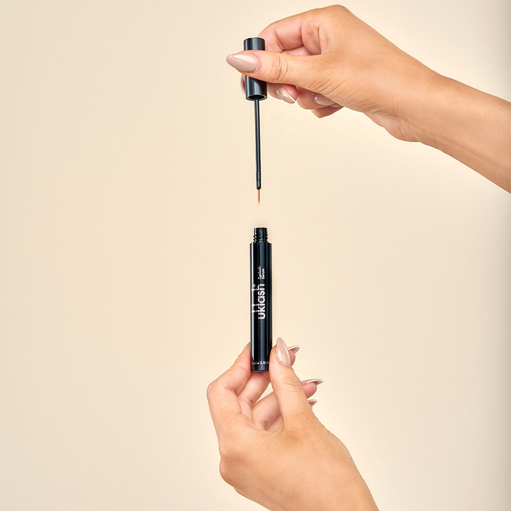 A person holds a UKLASH Eyelash Serum (3ml) container with its black cap, unscrewing it to reveal a slender brush. The formula is cruelty-free and promises longer eyelashes, set against a soft beige background.