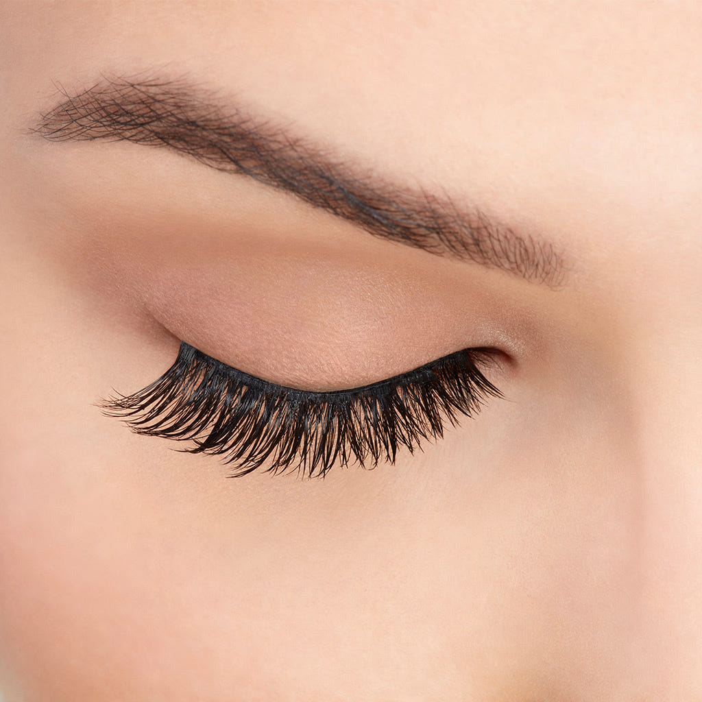 Close-up of a person's face with an eye closed, showcasing long, dark Ardell Faux Mink Lashes Black Demi Wispies and a neatly groomed eyebrow. The skin appears smooth and flawless, highlighting the natural beauty of the lashes.