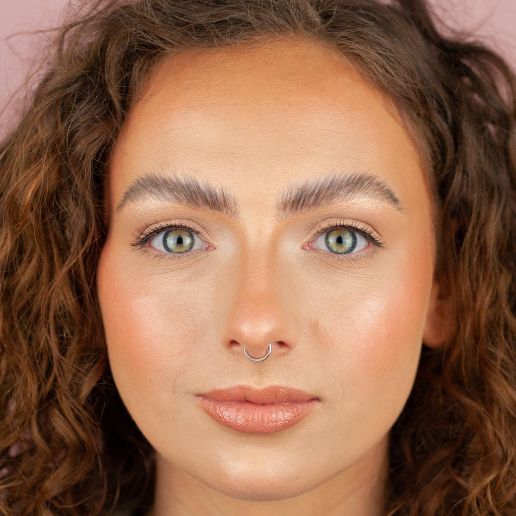 Close-up of a person with curly brown hair, light blue eyes, and a septum piercing looking directly at the camera. With distinct brows and Boldface Lashes - Short and Sweet, they're wearing cruelty-free natural makeup and a soft pink lip color.