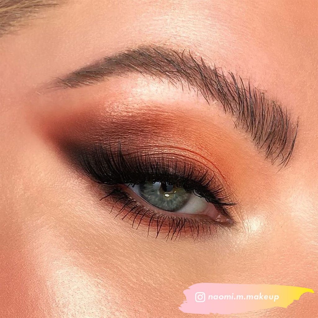 Close-up of an eye with striking makeup: featuring shimmery gold and deep copper eyeshadow, thick black eyeliner, and the voluminous Ardell Faux Mink Lashes in Black Demi Wispies. The eyebrow is meticulously groomed. The makeup artist's handle "naomi.m.makeup" is visible in the bottom right corner.